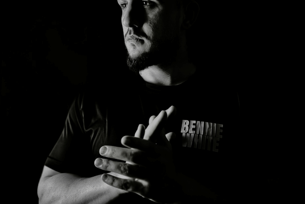 Black-and-white portrait of a bald, strong, masculine man standing in darkness with hands clasped, embodying presence, power, and purpose.