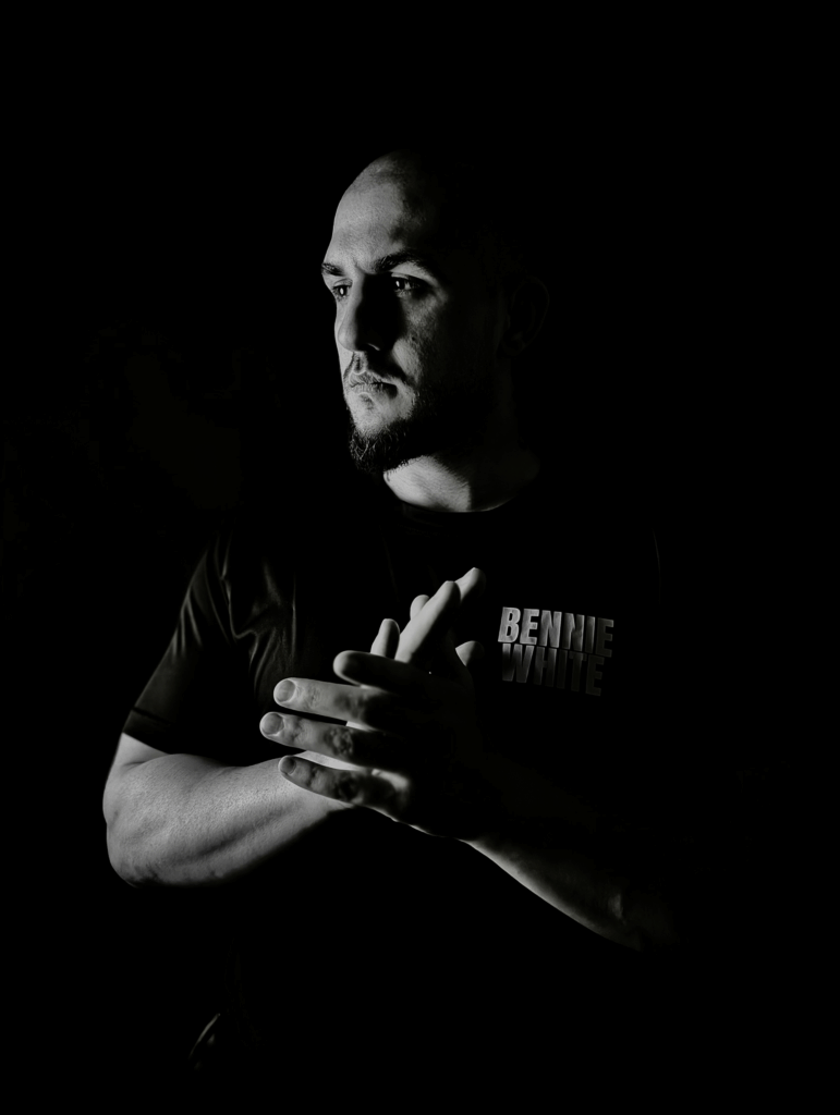 Black-and-white portrait of a bald, strong, masculine man standing in darkness with hands clasped, embodying presence, power, and purpose.